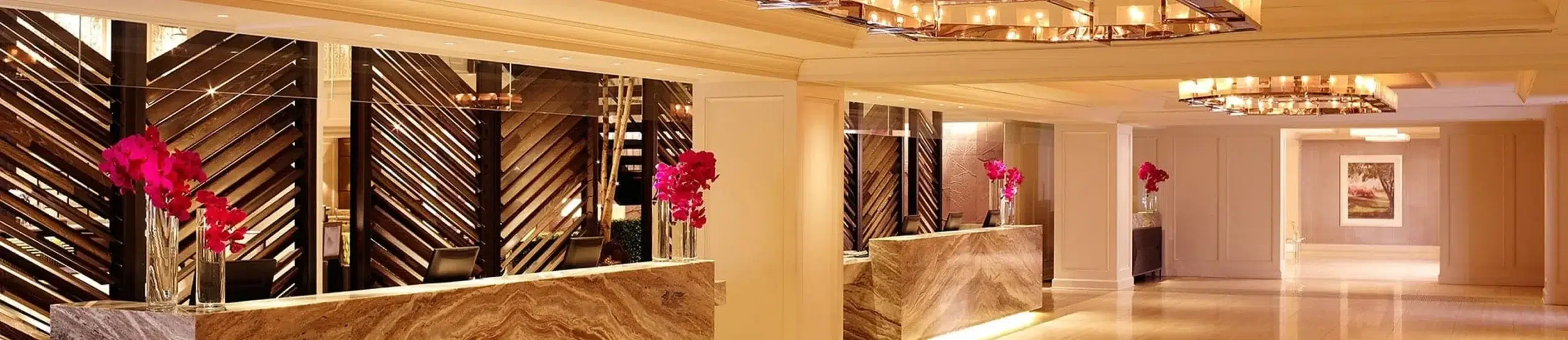 A luxurious hotel lobby with a marble reception desk, pink flowers, and a chandelier.