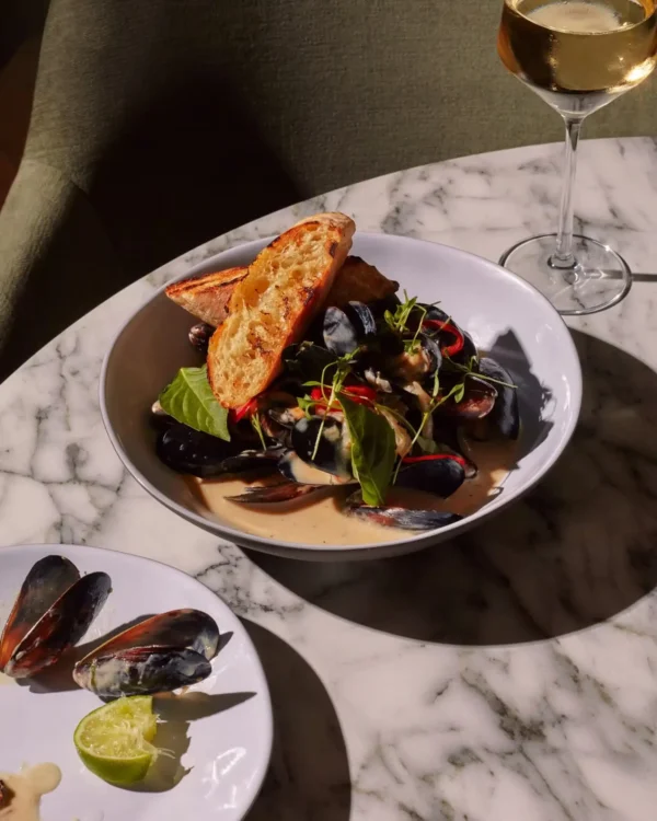 a bowl of mussels in a creamy sauce with bread and a glass of wine on a marble table