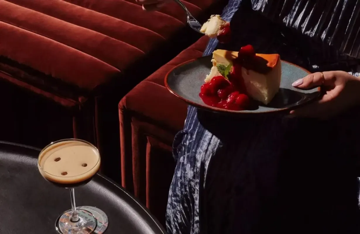 A plate of cheesecake with berries and a coffee drink on a table in a booth.
