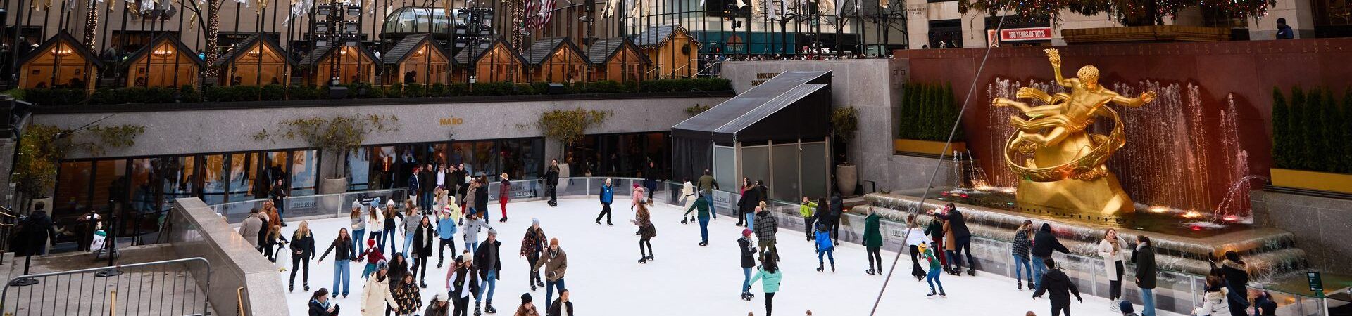 A large ice skating rink with people skating, a fountain, and a statue in front of a building.